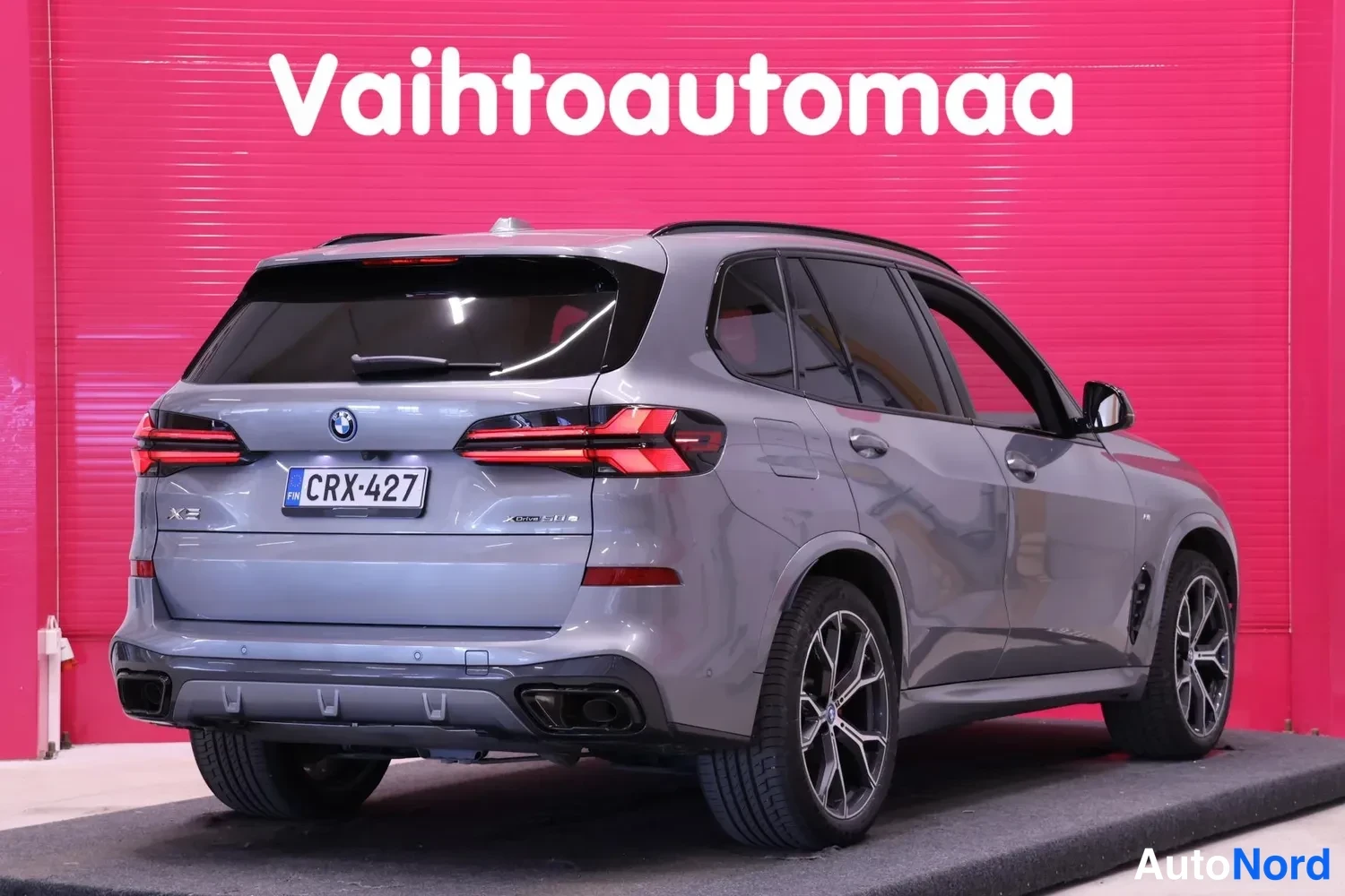Takaviisto: Auton takanurkkakuva: harmaa BMW X5 takaa vasemmalta kuvattuna, rekisteri, taustana punainen Vaihtoautomaata-logo.