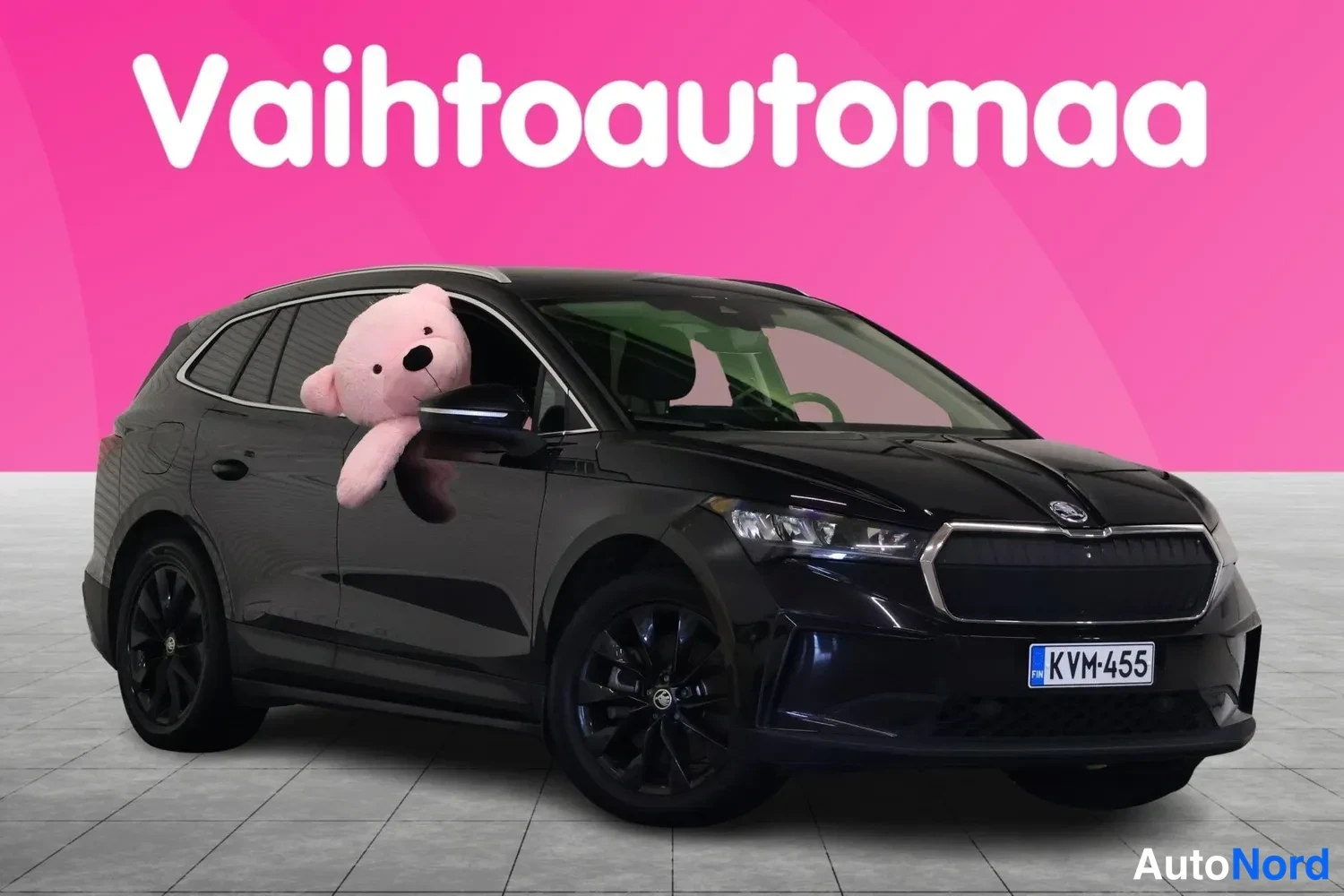 Etuviisto, nokka oikealle: Mustan tilavautomobiilin etukulmanäkuva rekisterikilpi, pinkki tausta tekstillä Vaihtoautomaa.