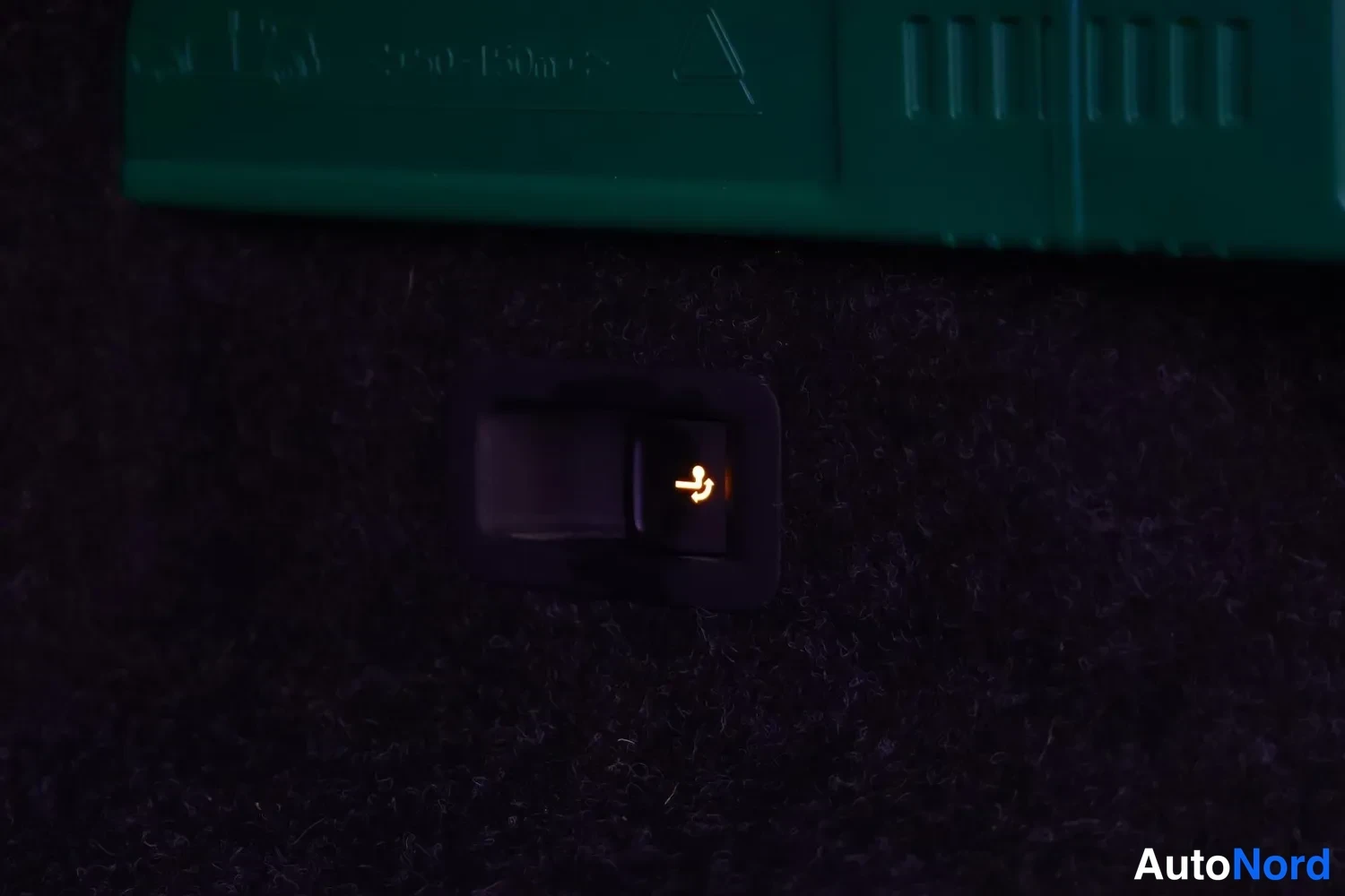 Yksityiskohta: Illuminated orange ISOFIX anchor symbol indicator switch on dark carpet floor next to green storage tray