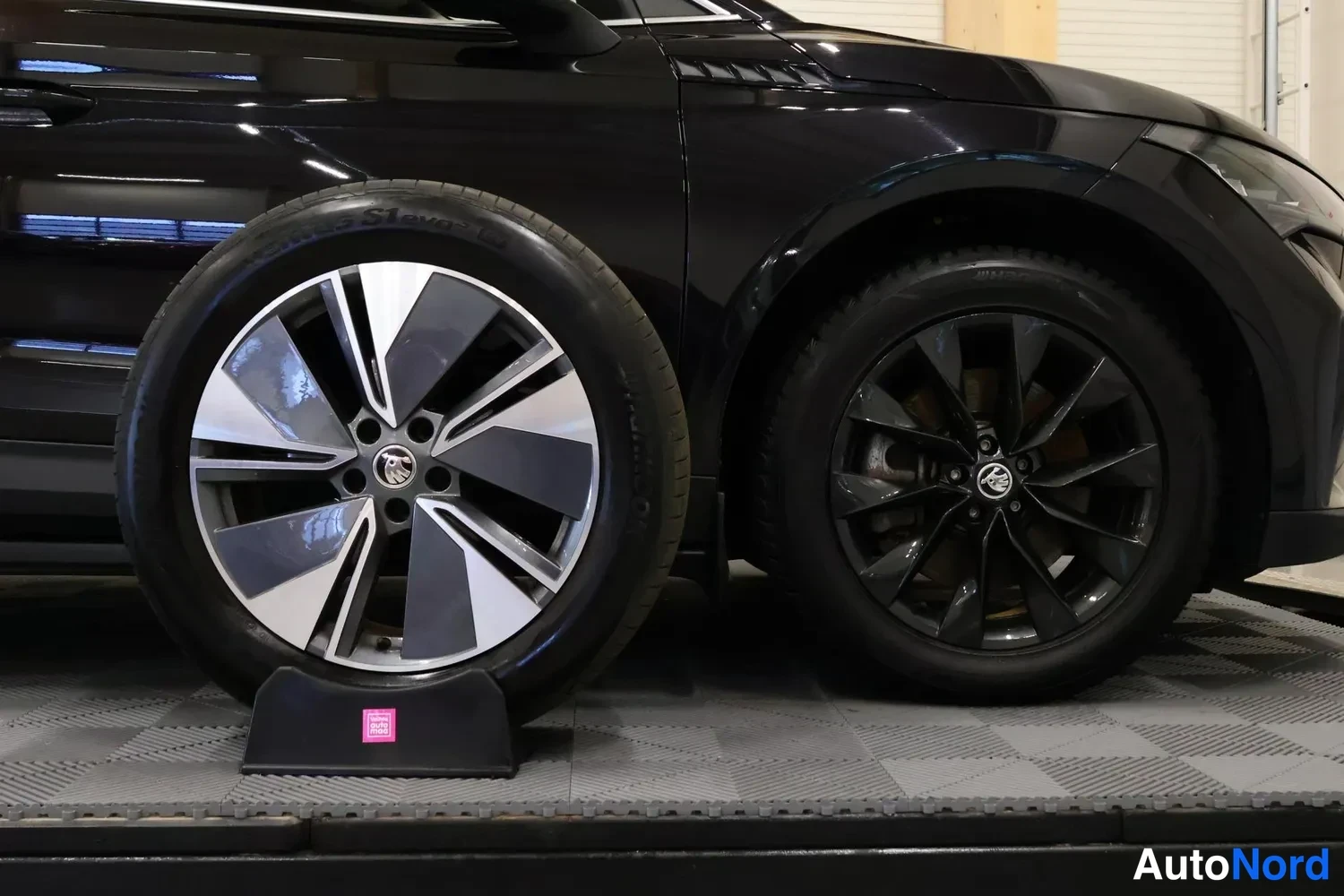 Sivulta: Side profile of black vehicle showing front and rear wheel arches; front wheel features two-tone alloy rim resting on black chock while rear wheel is solid blac