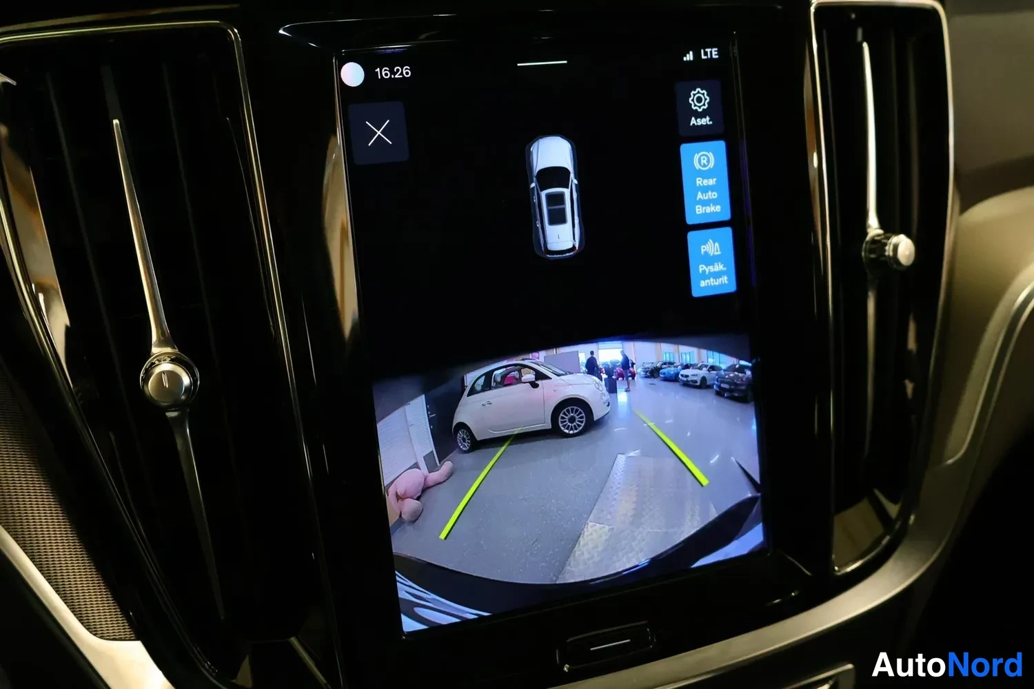 Yksityiskohta: Interior shot of vehicle rearview camera display on central infotainment screen. Shows live view through wide-angle lens of parking garage floor with yellow gui