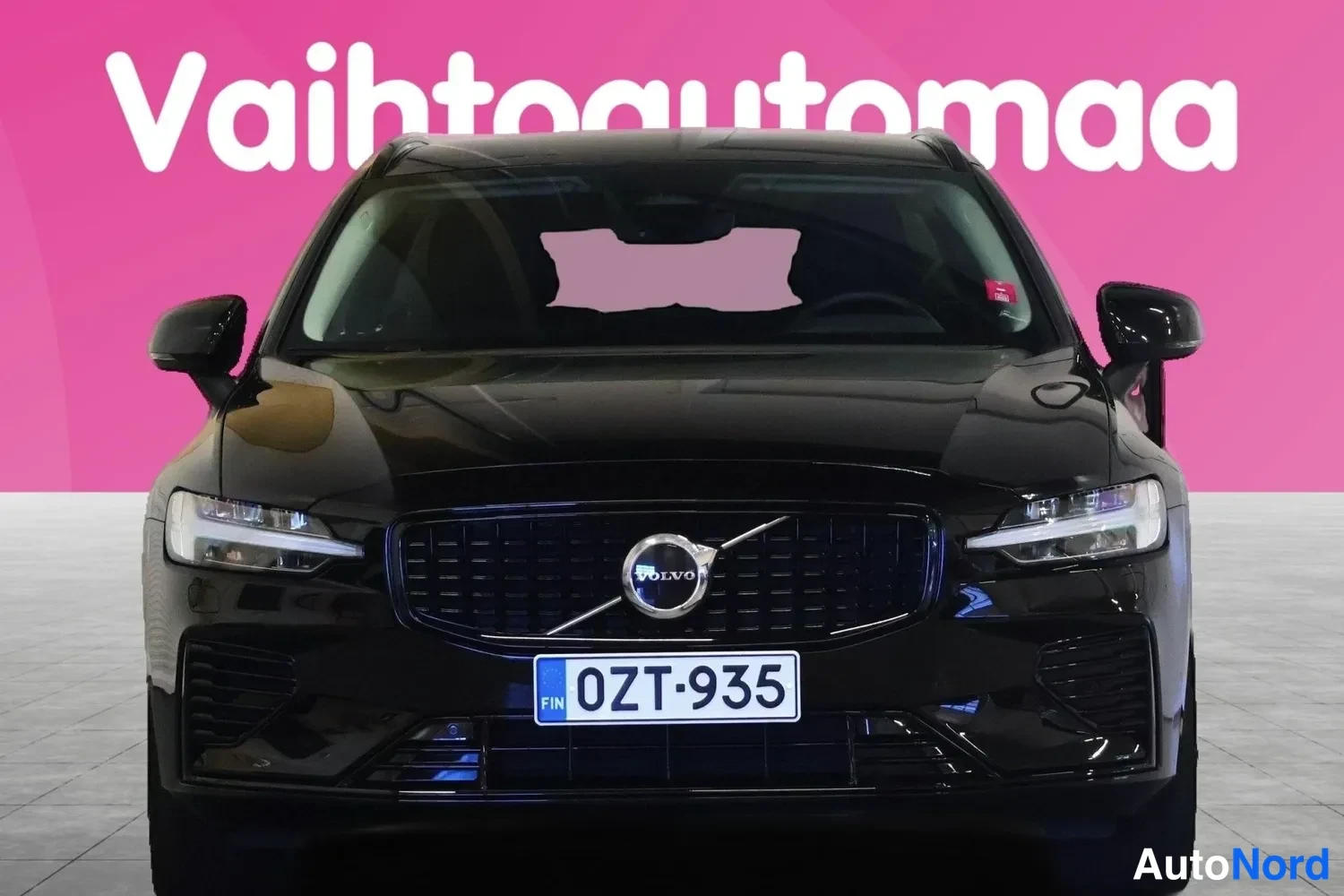 Edestä: Musta farmariauto suoraan edestä kuvattuna, Volvo-merkki kylpyläritilässä, rekisteritunnus.