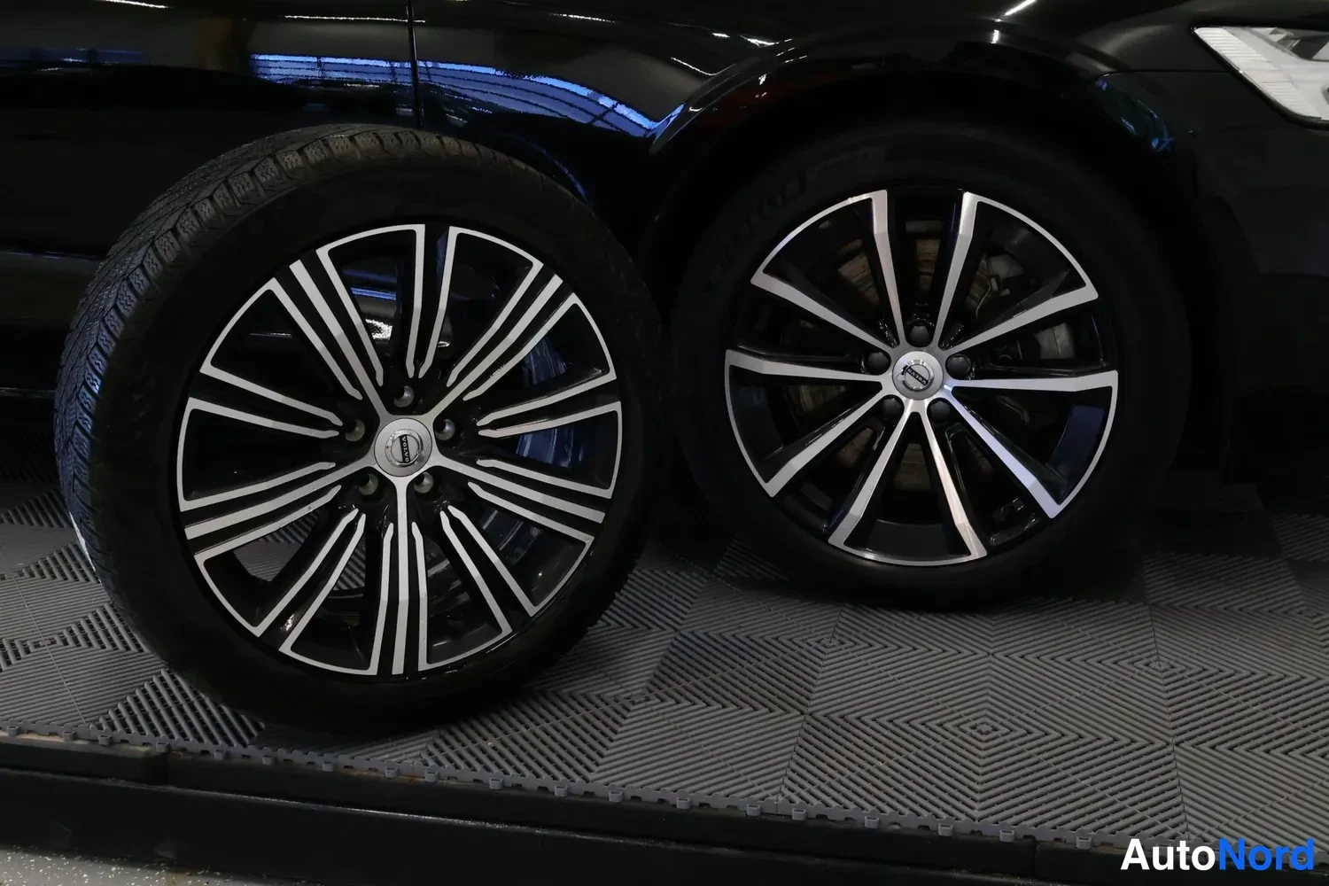 Muu: Kaksi Volvo-kestovaappaan pölyä kimaltelevaa kevytmetallivannetta; vasemmalla pölyä ja oikealla auton pyörä; musta karjakuviollinen lattia.
