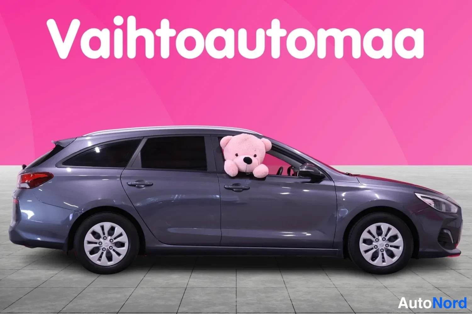 Etuviisto, nokka oikealle: Hyundai i30 -farmari colottu autotarvikeliikkeen mainospohjalla; tummanharmaa korirakenne, etusälekaitti ja oikeanpuoleinen kylki näkyvissä; pehmolelu au