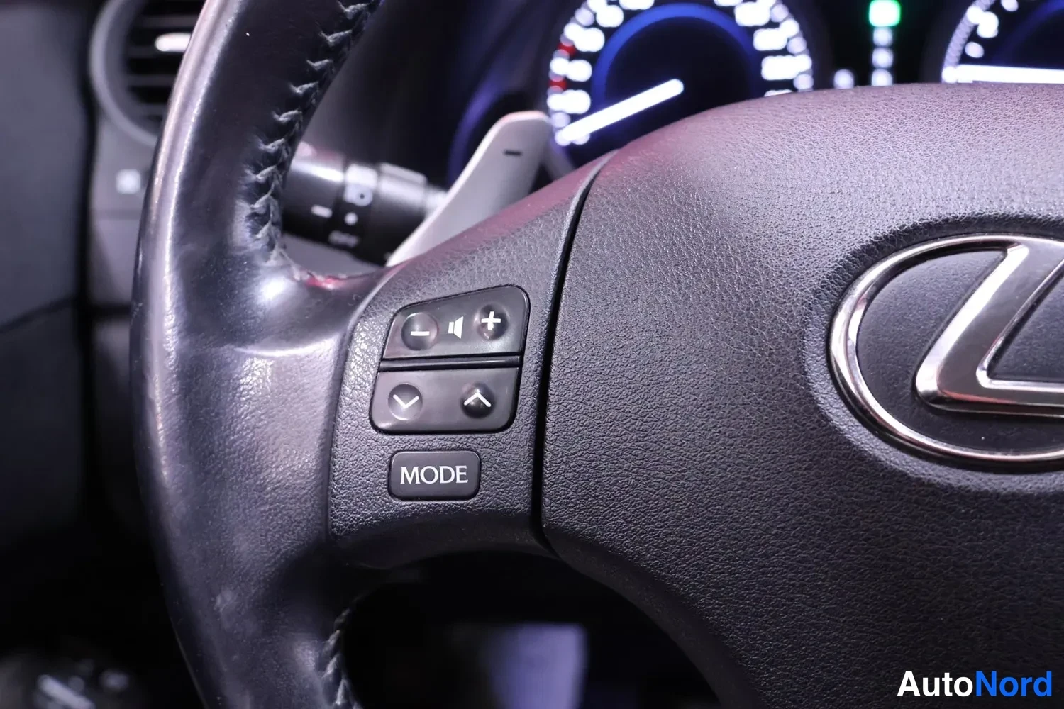Yksityiskohta: Ohjauspyörän vasemman puolen levykän näkymä: äänenvoimakkuus- ja äänenpaine +/- ja nuolipainikkeet, MODE-tekstillä varustettu painike, Lexus-logo oikealla.
