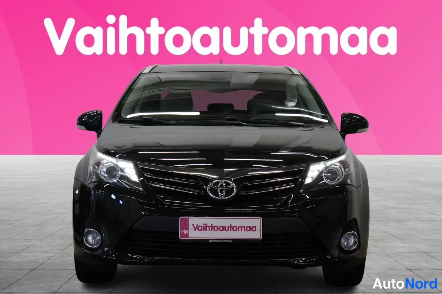 Edestä: Musta Toyota Avensis -farmari suoraan edestä, Toyota-merkki ja sumuvalot näkyvissä.