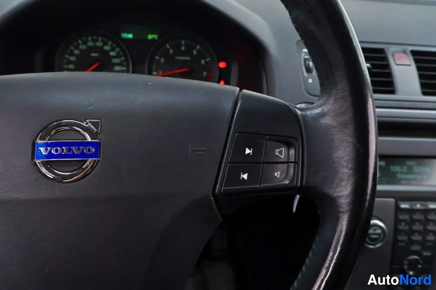 Muu: Volvo C30:n ohjauspyörä lähikuvassa; Volvo-emblem ja äänentoiston ohjainpainikkeet näkyvät, taustalla osa mittaristosta ja kojelaudasta.