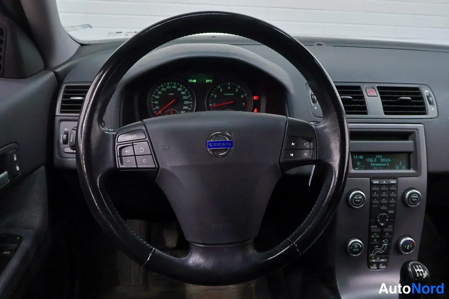 Muu: Kuljettajan näkymä Volvo C30:n kojelautaan: ohjauspyörä cruise-painikkeilla, mittaristo, radio-näyttö tekstillä 'FM1 105.2 NOVA' ja manuaalivaihteiston vaihdeke