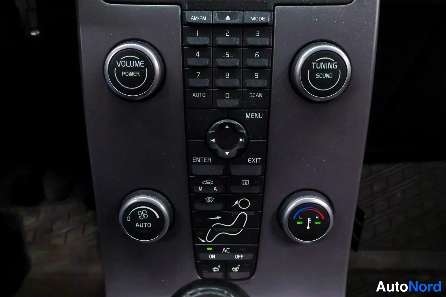 Muu: Volvo C30:n keskiöidyn kojelaudan keskusyksikkö lähikuvassa: num Näppäimistö, VOLUME/TUNING-kehrä, AUTO-ilmastointipainike ja lämpötilanäyttö L/R; AC ON/OFF-tek