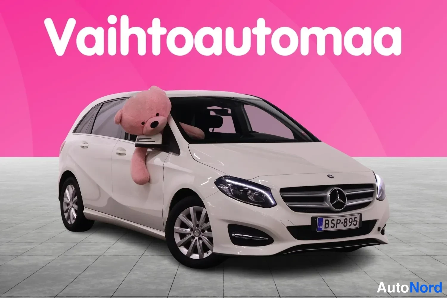 Etuviisto, nokka oikealle: Valkoinen Mercedes-Benz B-sarjan farmariauto kolmioneljännes etunäkymä, keulakuva oikealla, suuri pehmolelu tuijottamassa ikkunasta.