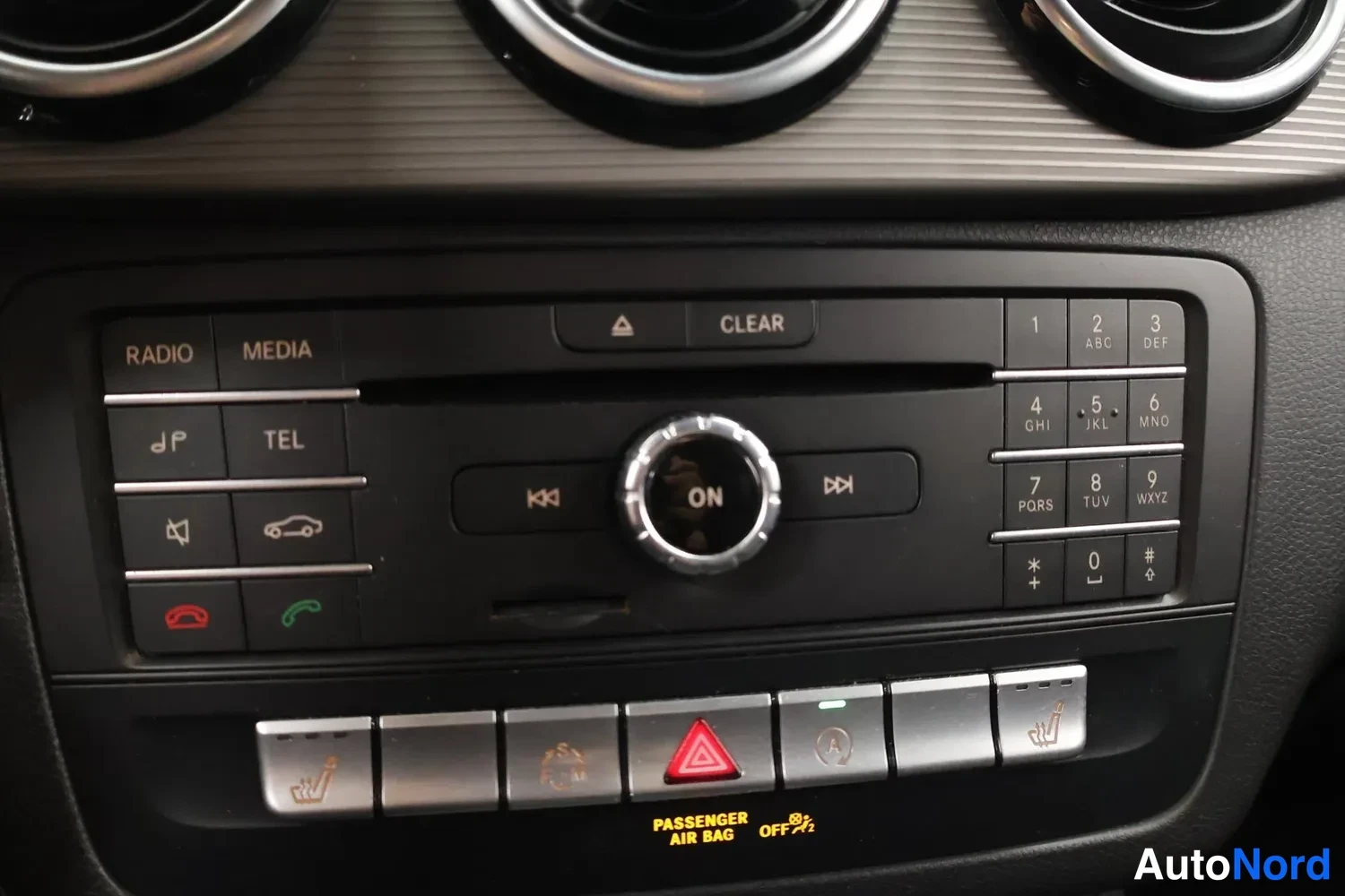 Yksityiskohta: Keskikonsolin painikkeet: RADIO, MEDIA, TEL, CD-ahto, numeroitnäppäimistö ja varoitusvilkkujen nappi, alapuolella 'PASSENGER AIR BAG OFF'-merkintä.