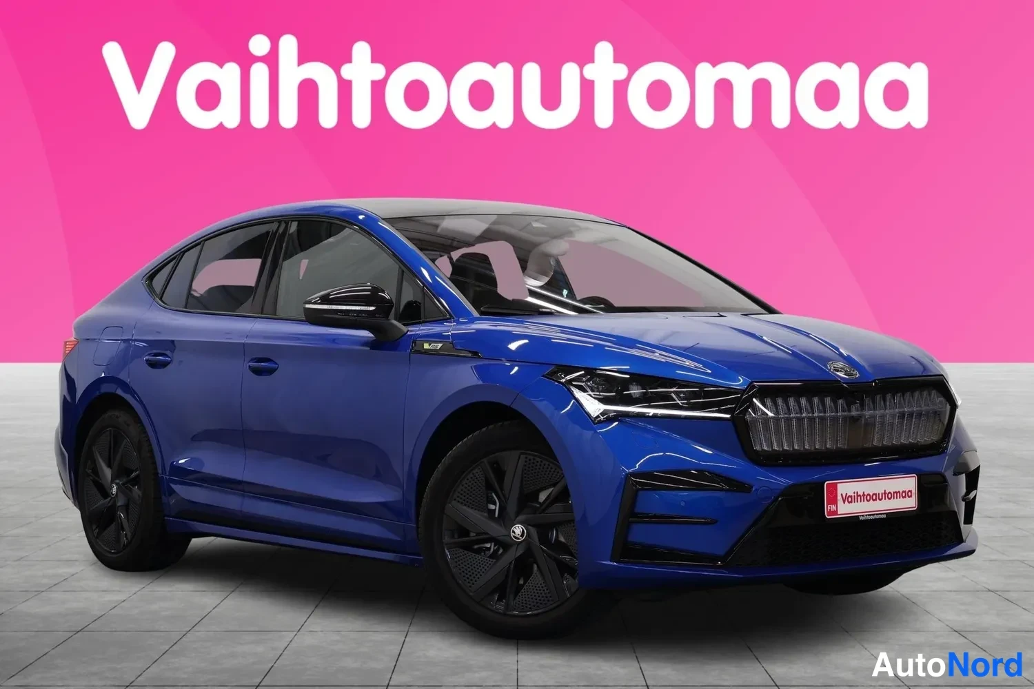 Etuviisto, nokka oikealle: Sininen Škoda Enyaq Coupé etuokolukuva oikealta, etupuskuri ja kylki näkyvissä pinkkitaustaa vasten.