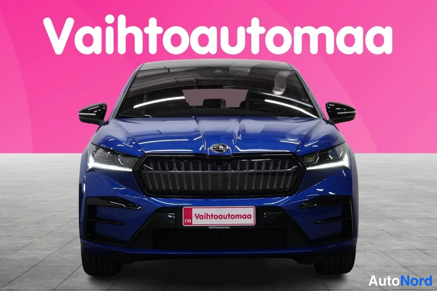 Edestä: Sininen Škoda Enyaq Coupé suoraan edestä, etusäleikkö ja ajovalot näkyvissä pinkkitaustaa vasten.