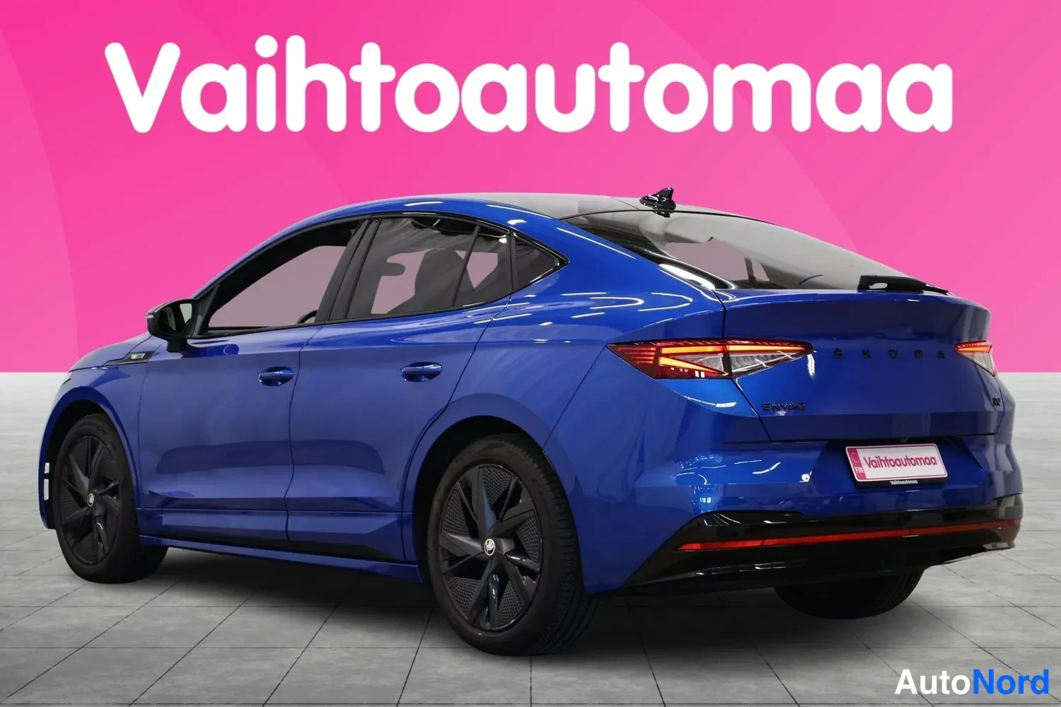 Takaviisto: Sininen Škoda Enyaq Coupé takaokolukuva vasemmalta, takavaloilla ja kylki näkyvissä pinkkitaustaa vasten.