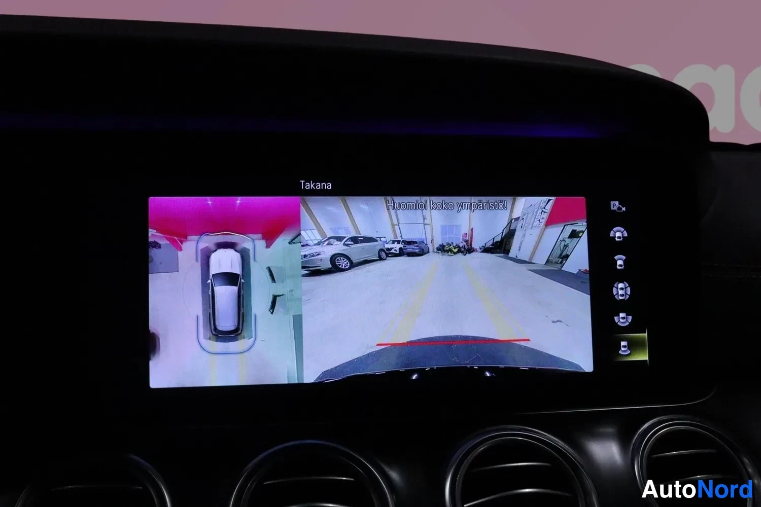 Yksityiskohta: Kojelaudan infotainment-näyttö, joka näyttää 360-asteen ympäristökameran kuvan: vasemmalla ylhäältä kuvattu ajoneuvo ja oikealla takakameran näkymä tilasta, jos