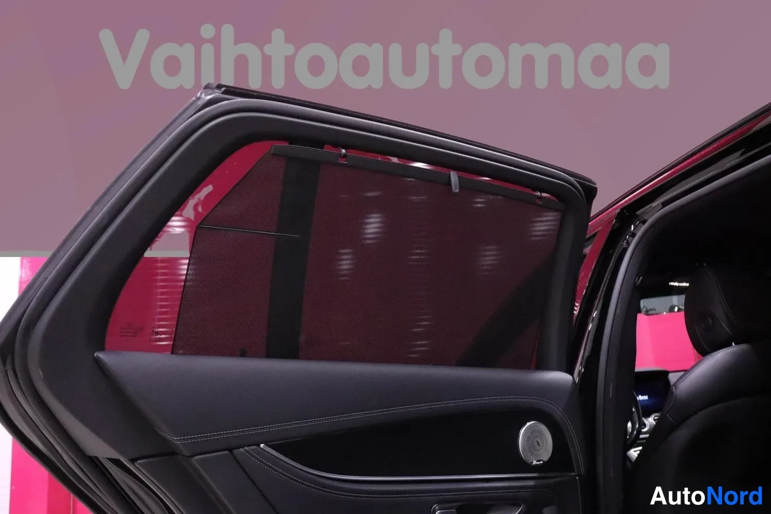 Yksityiskohta: Avattu takaovi sisäpuolelta: tumma sivuikkunasuoja vedettynä ikkunaan ja musta ovipaneeli kaiuttimeen, taustalla pinkki Vaihtoautomaa-tausta.