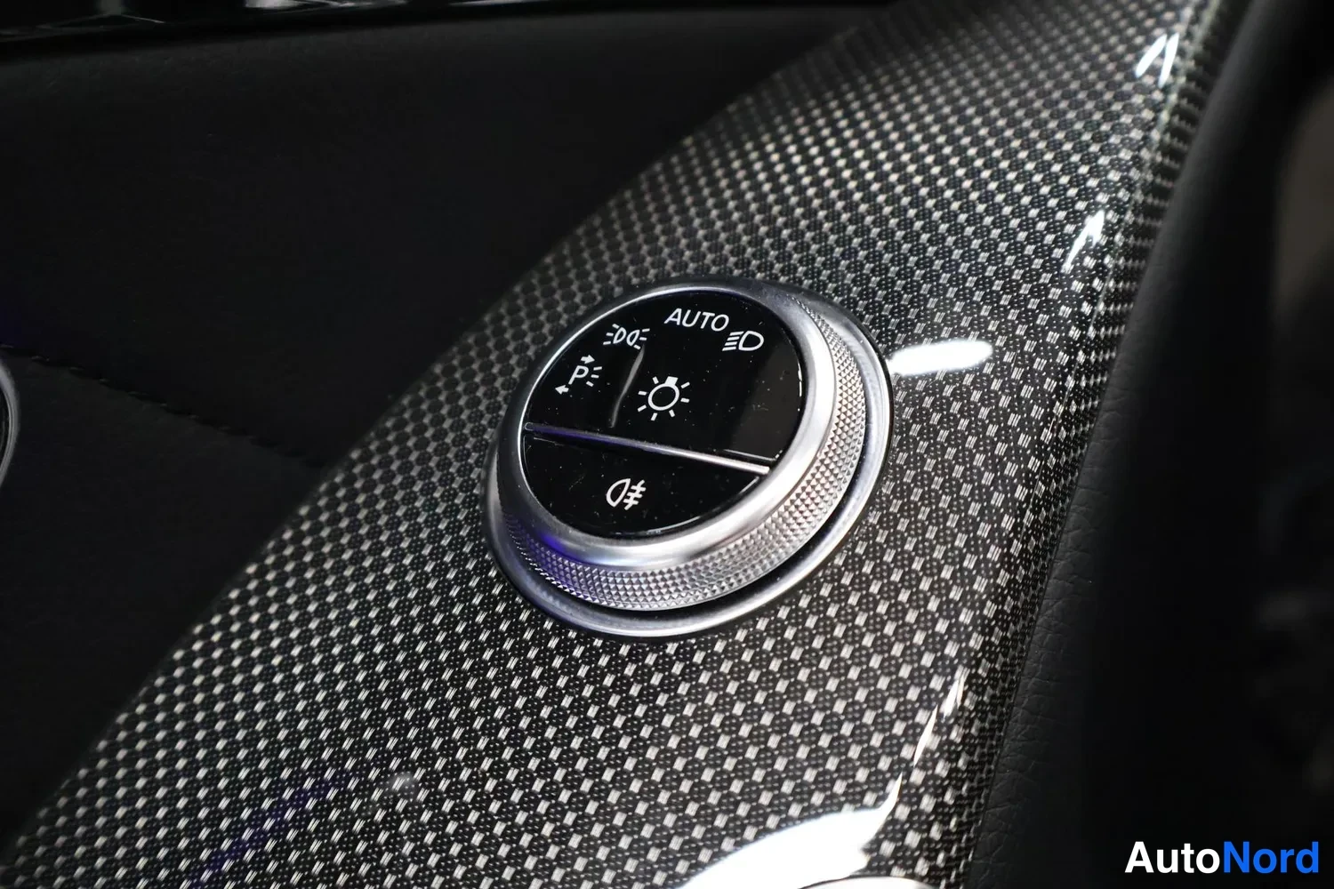 Muu: Sisätilakuva: pyöreä valaistuksen hallintakytkin, jonka merkinnöissä lukee AUTO, hiilikuitukuvioisen koristepinnan keskellä.