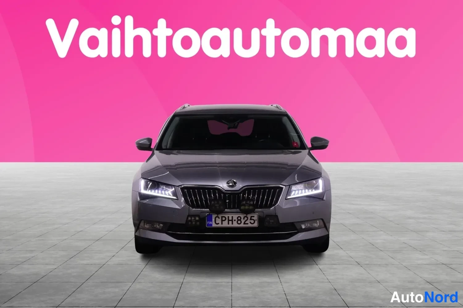 Edestä: Harmaa farmariauto suoraan edestä, rekisteritunnus, etuvalot päällä vaaleanpunaisella taustalla.