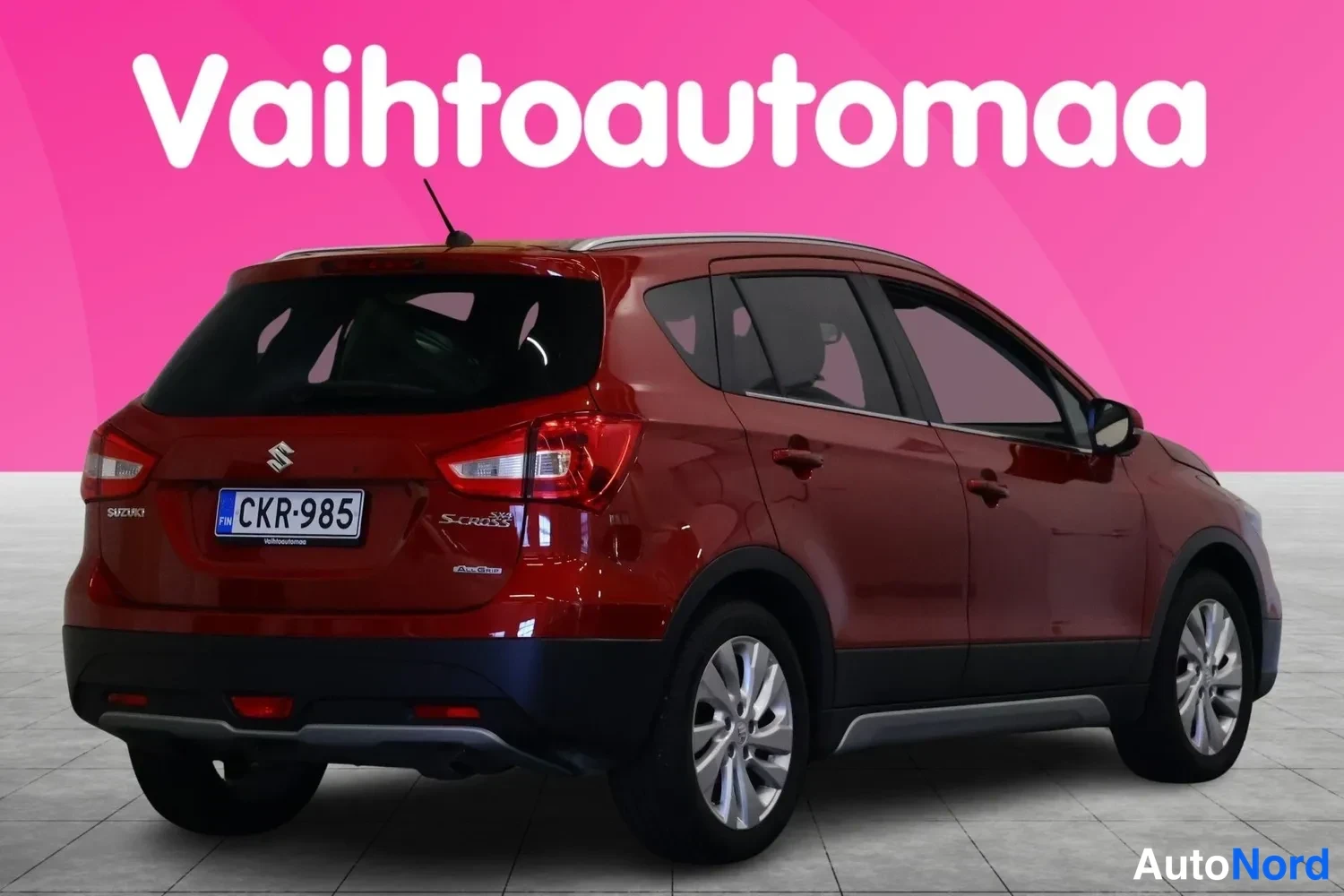 Takaviisto: Punainen tila-auto takaa ja oikealta sivulta nähtynä, rekisterikilpi, S-Cross-merkintä, pinkki tausta.