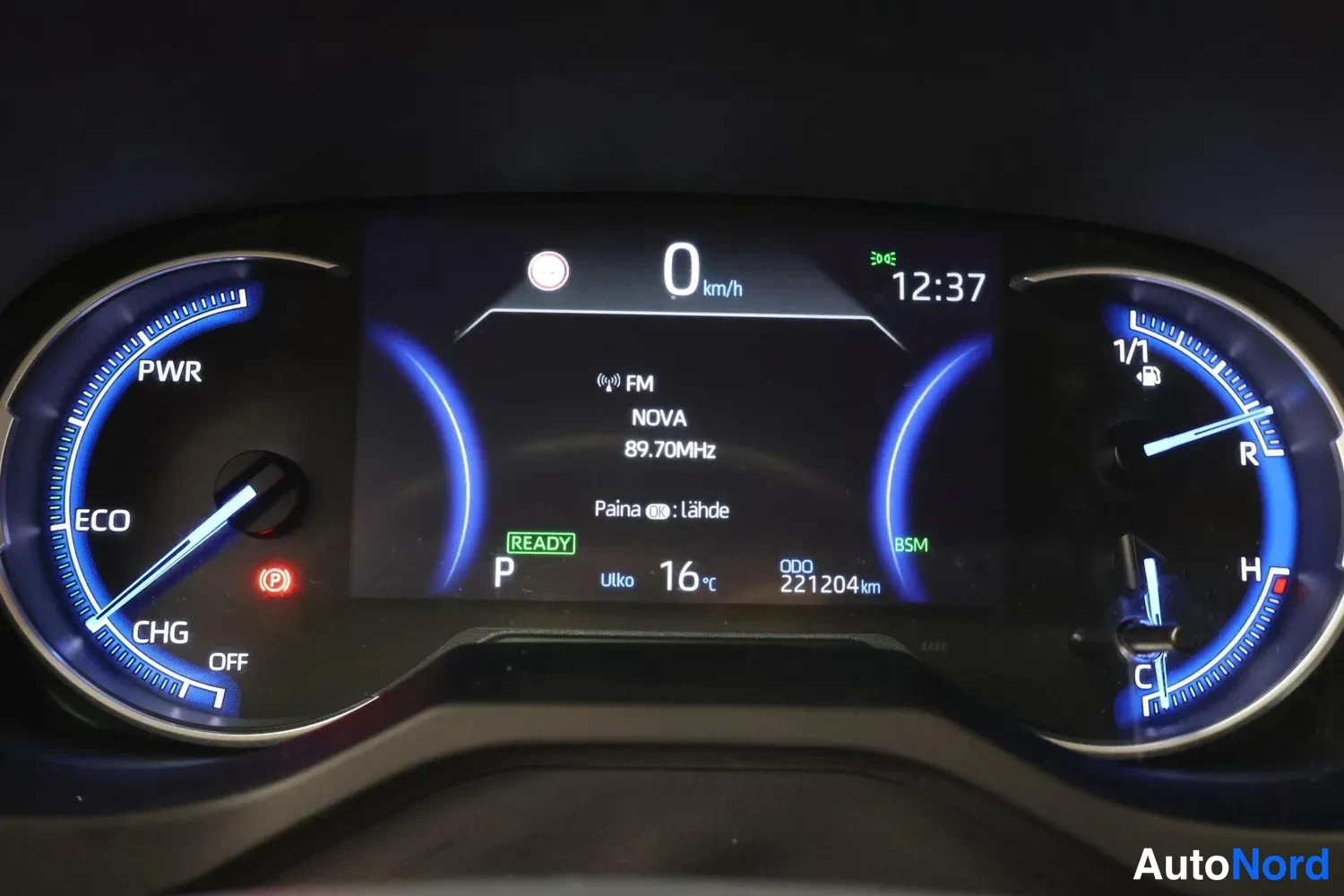 Muu: Mittaristoston näyttö: hybridiauto READY-tilassa, P-vaihde, nopeus 0 km/h, kokonaislukema 221204 km, radio Nova 89.70 MHz, ulkolämpötila 16 °C, kello 12:37, PWR