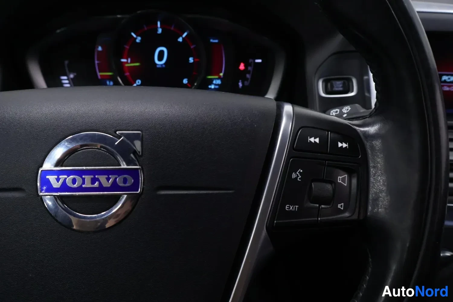 Yksityiskohta: Ohjauspyörän Volvo-merkki ja volyymi- ja ääniohjaimet, taustalla mittaristo.