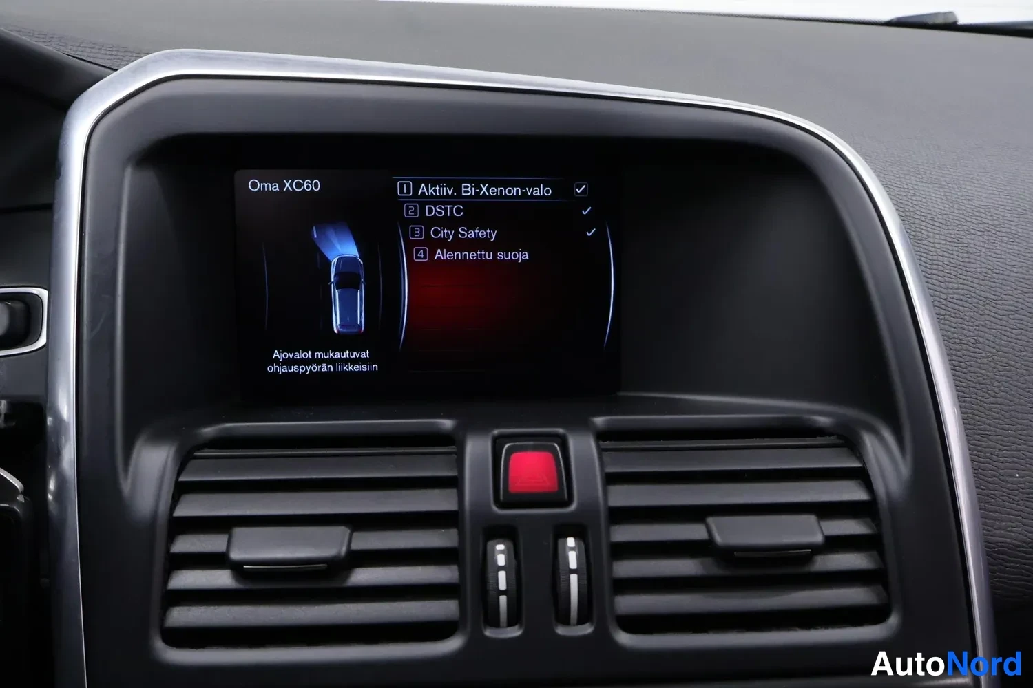 Yksityiskohta: Kojetaulun näyttö, jossa lukee 'Oma XC60' ja lista: 'Aktiv. Bi-Xenon-valo', 'DSTC', 'City Safety', 'Alennettu suoja'. Näytön alla ilmastointisuuttimet ja punain