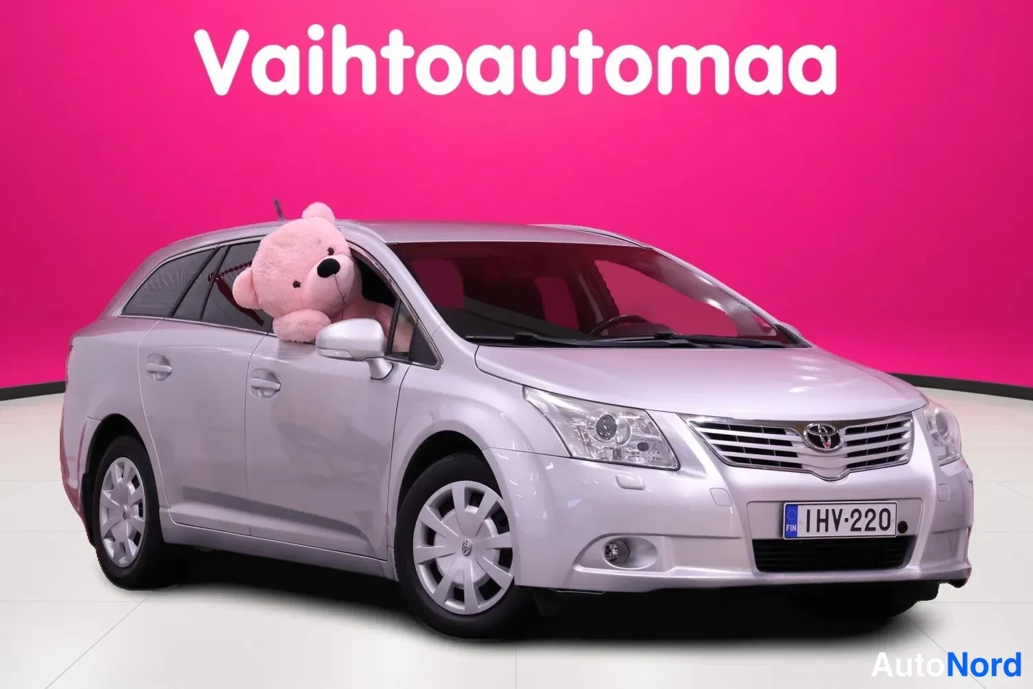 Etuviisto, nokka oikealle: Hopeanharmaa farmariauto kolmannesoveltuvassa etunäkymässä, nokka oikealla, valkoinen teddykarhu kuljettajan ikkunasta ulkona, taustalla vaaleanpunainen