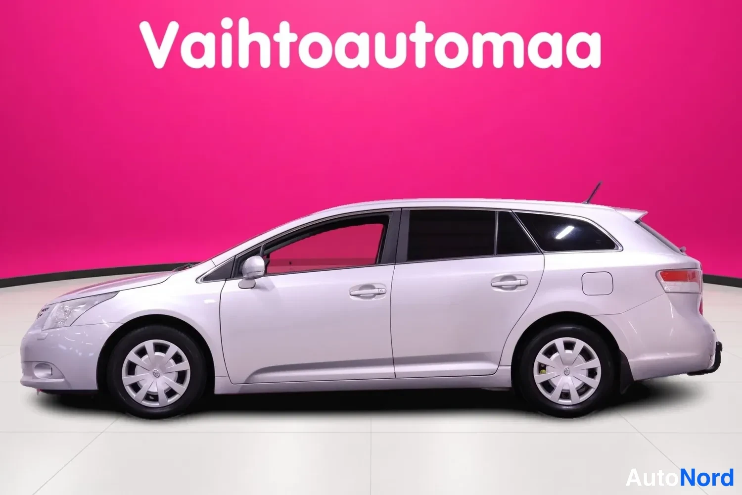 Etuviisto, nokka vasemmalle: Hopeanharmaa farmariauto sivusuunnassa vasemmasta etukulmasta valot studion vaaleanpunaista taustaa vasten.