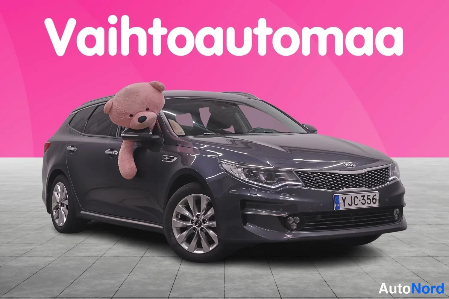 Etuviisto, nokka oikealle: Kia Optima -farmari tummanharmaana etukulmasta rekisteritunnus, teddykarhu ikkunassa