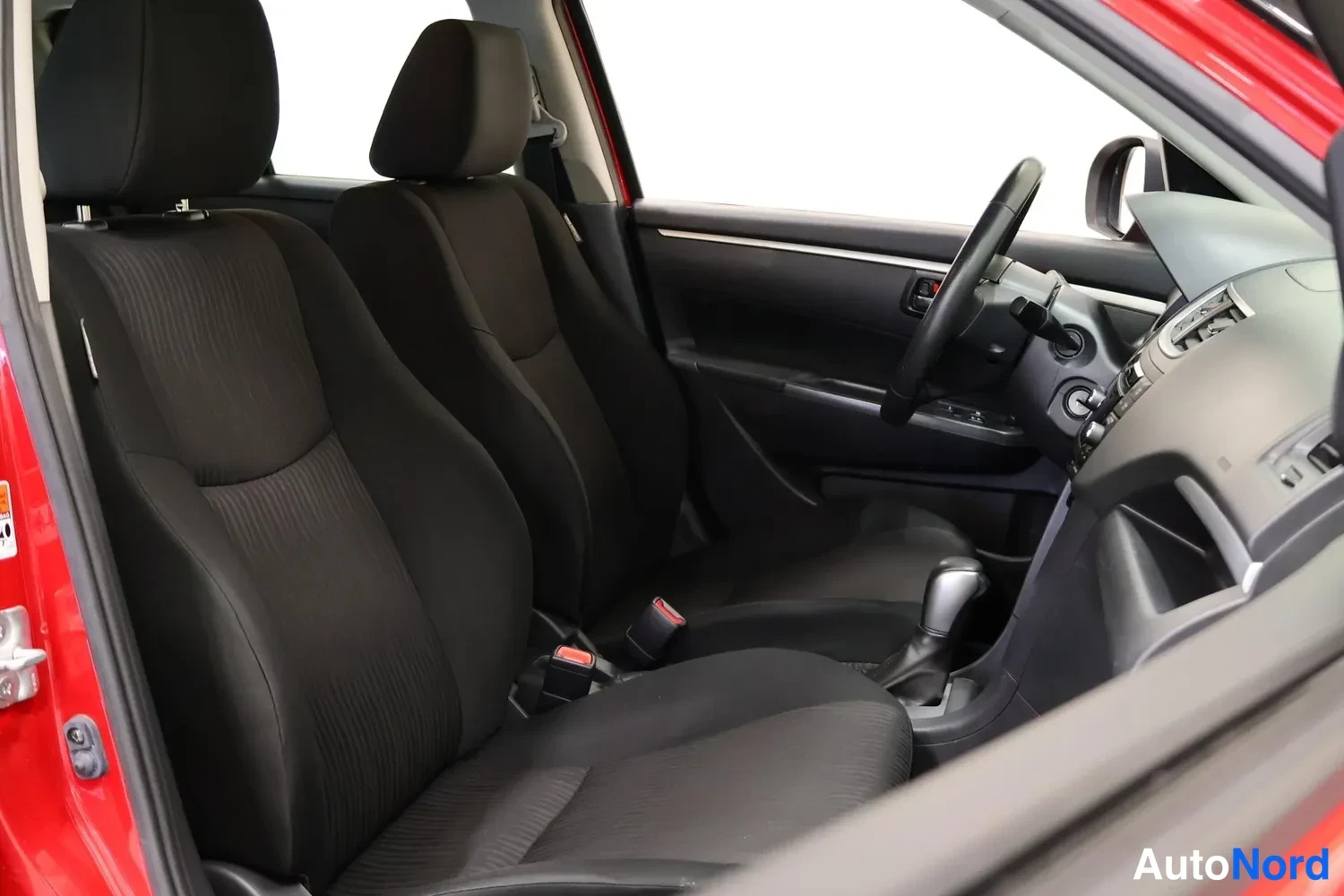 Muu: Suzuki Swiftin etusija ulkoa kuvattuna: kaksi mustaa kudintoa etuistuinta, ohjauspyörä oikealla ja osa kojelautaa näkyvät.