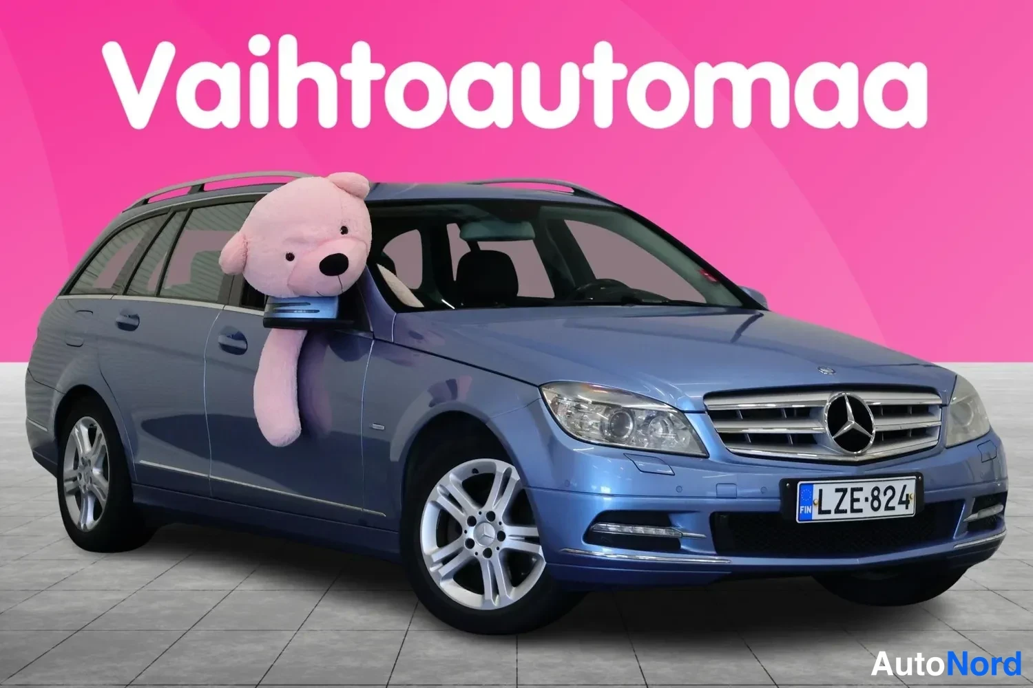 Etuviisto, nokka oikealle: Sininen Mercedes-Benz farmari kolmannesovelta kuvattuna, tunnus, vaaleanpunaisella taustalla.