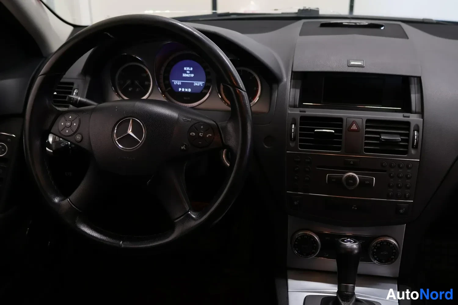 Muu: Kuljettajan puolen näkymä Mercedes-Benzin ohjaamoon: ohjauspyörä, mittaristo ja keskikonsoli, vaihteenvalitsimen näytössä 'PRND'.