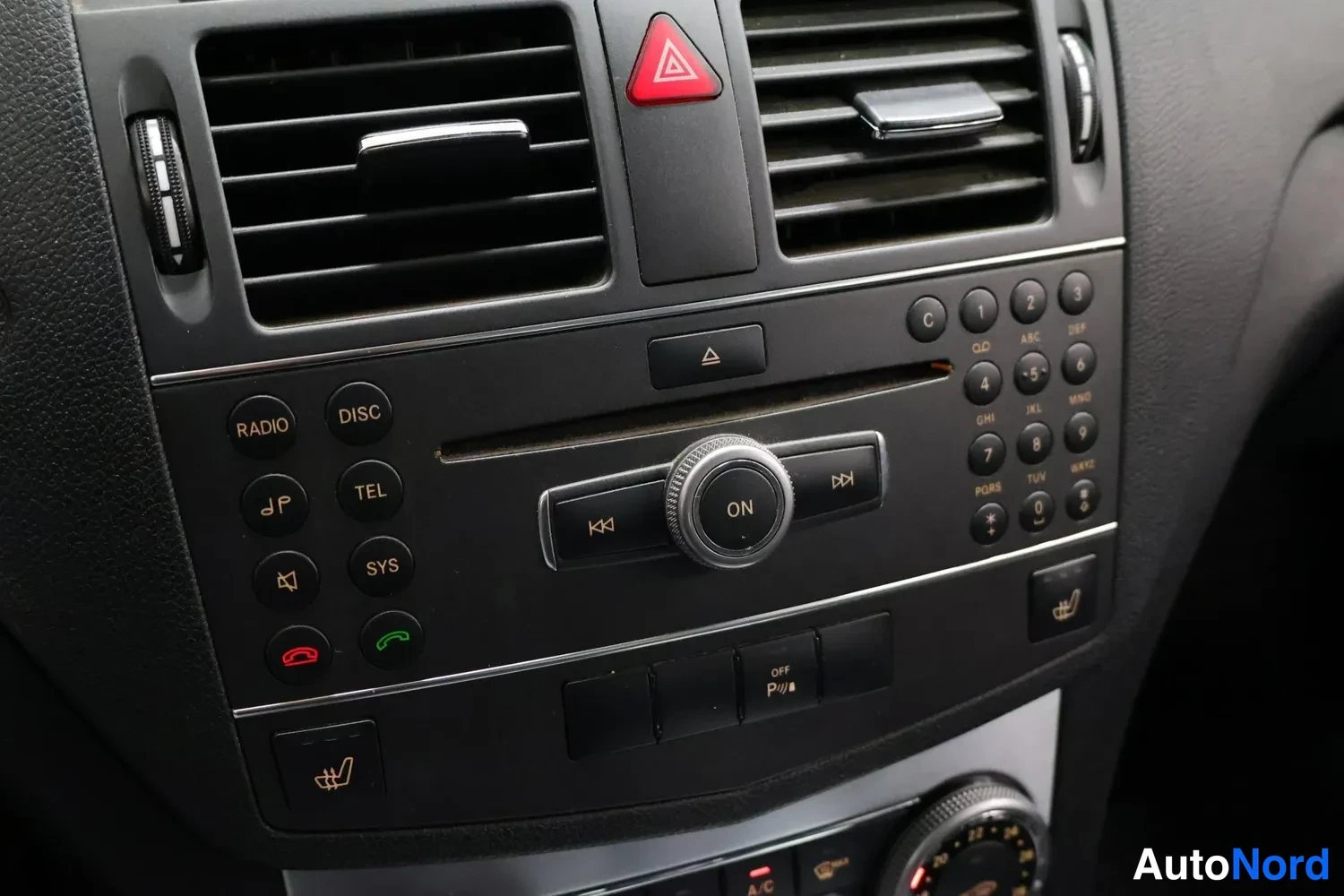 Yksityiskohta: Sisätilakuva Mercedes-Benz C 200 d 2011:n keskikonsolista: kaksi ilmanvaihtoaukkoa, punainen varoitusvalokolmio, radio/CD-yksikkö, ON-kiertosäädin, puhelin- ja
