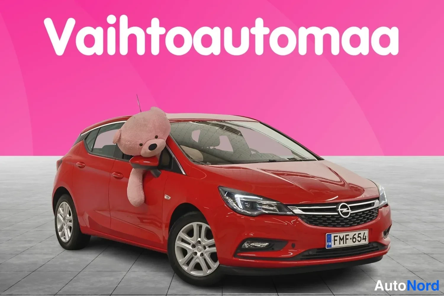 Etuviisto, nokka oikealle: Punaisen Opel Astran etukulmanotto, keula ja vasen kylki näkyvissä; etupuskurin rekisterikilpi ja suuri pehmolelu sivupeilin kohdalla.