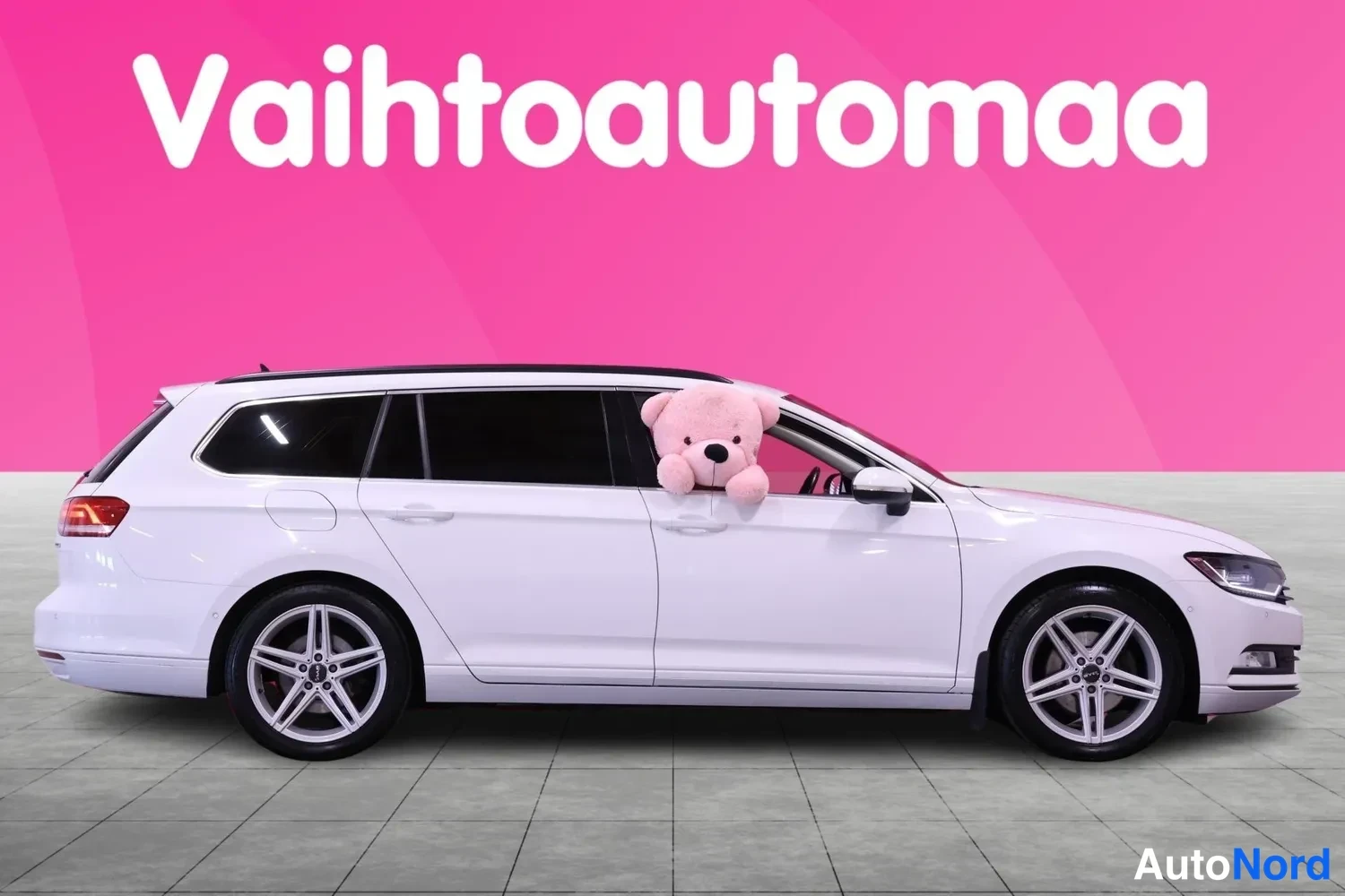 Etuviisto, nokka oikealle: Valkoinen farmariauto sivusta‑etukulmasta, keula oikealla, vaaleanpunaisella taustalla.