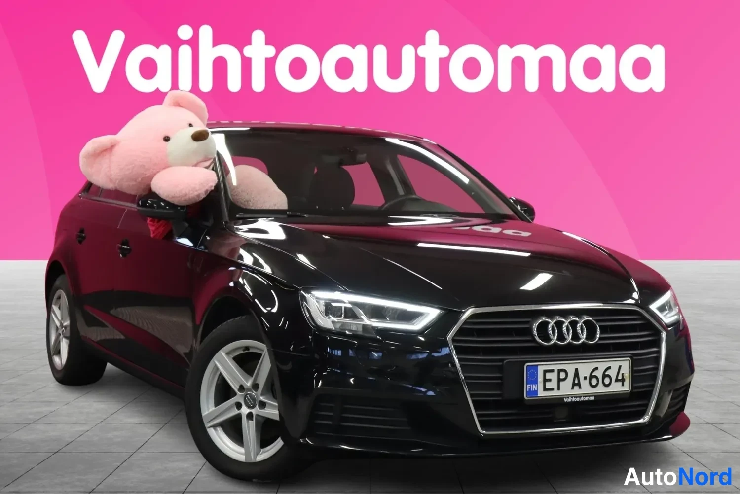 Etuviisto, nokka oikealle: Musta Audi A3 Sportback kolmesta neljäsosasta edestä oikealta, rekisterikilpi, pinkillä taustalla.