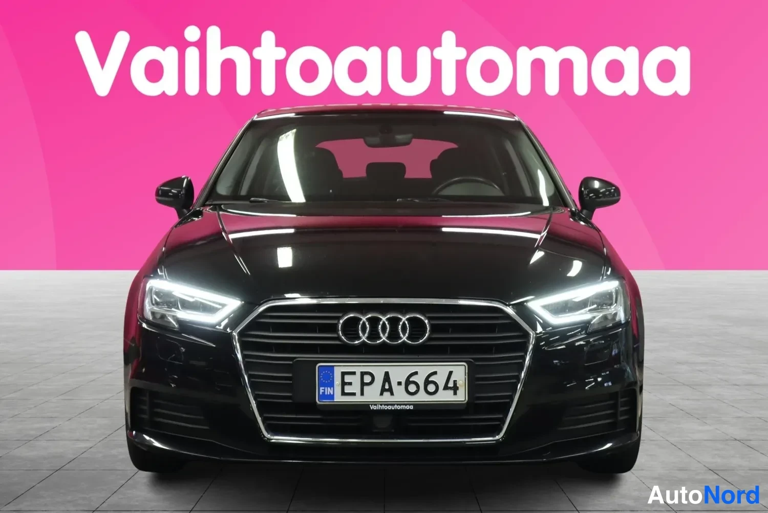 Edestä: Mustan Audi A3 Sportbackin etupää suoraan edestä, rekisterikilpi, pinkillä taustalla.