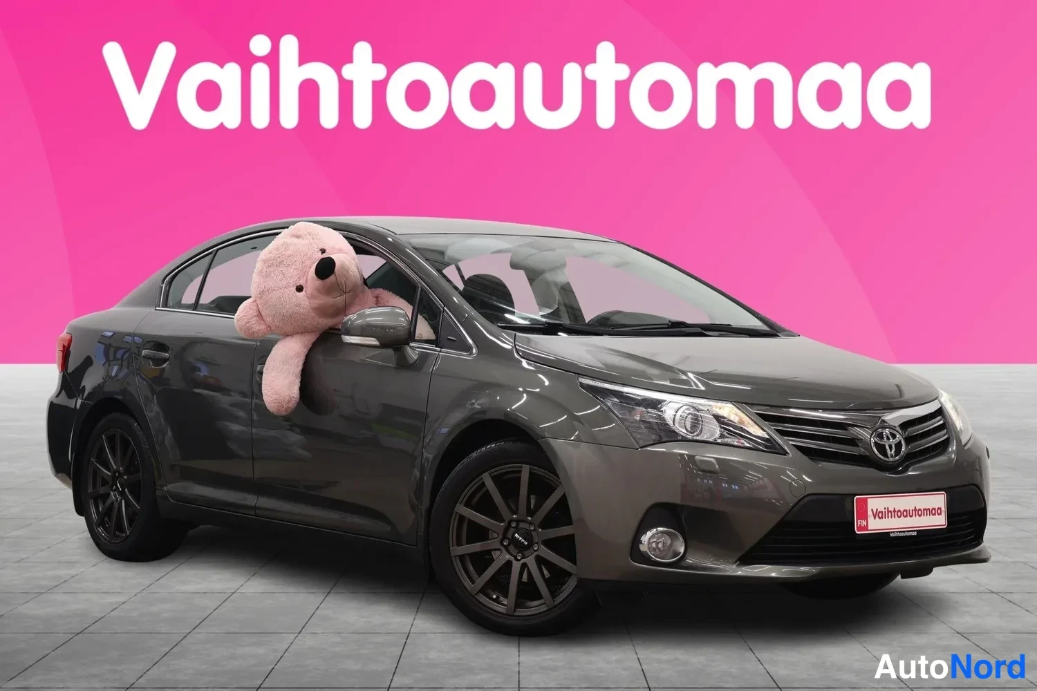 Etuviisto, nokka oikealle: Auton etukulma ja vasen sivu; keula oikealla, harmaanruskea sedan, tummat kevytmetallivanteet, pehmolelu takasivuikkunassa.
