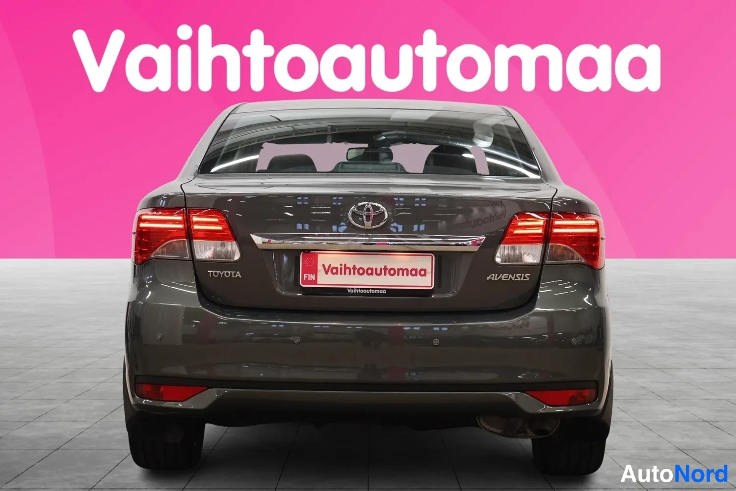Takaa: Auto suoraan takaa kuvattuna: Toyotan merkki, AVENSIS-teksti ja Vaihtoautomaan rekkinumeron suojus takaluukussa, punaiset takavalot.