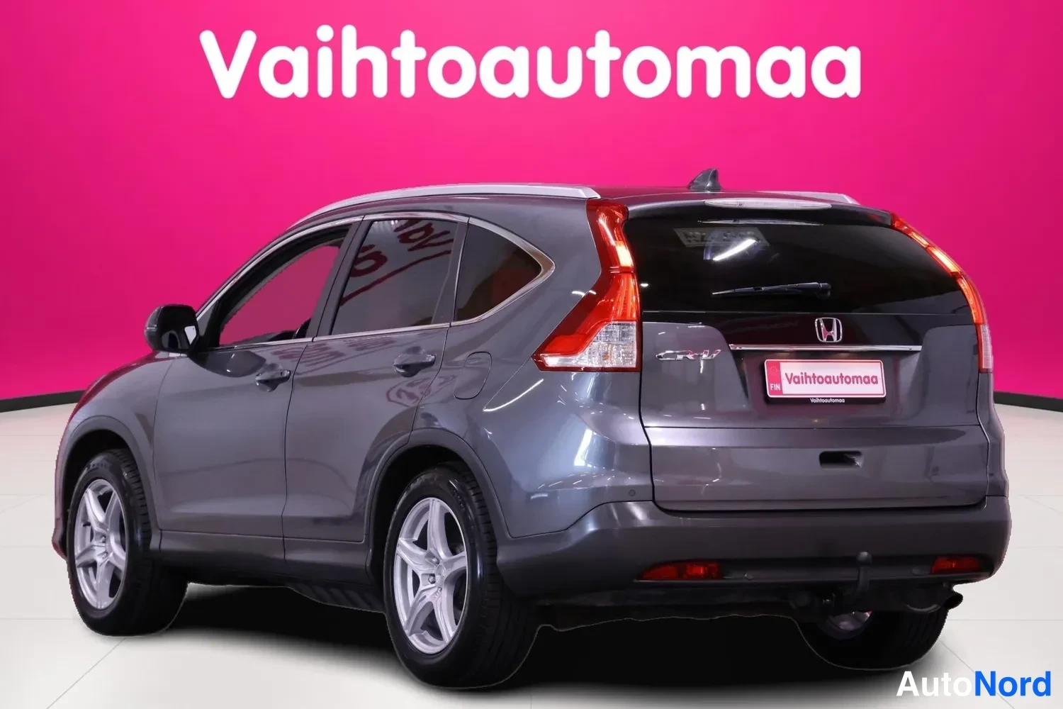 Takaviisto: Tummanharmaa Honda CR-V kuvattuna takaviistosta vasemmalta: takavalojen välissä Honda-merkki, katolla hopeiset kattokaiteet, pinkin taustan ja vaalean lattian e