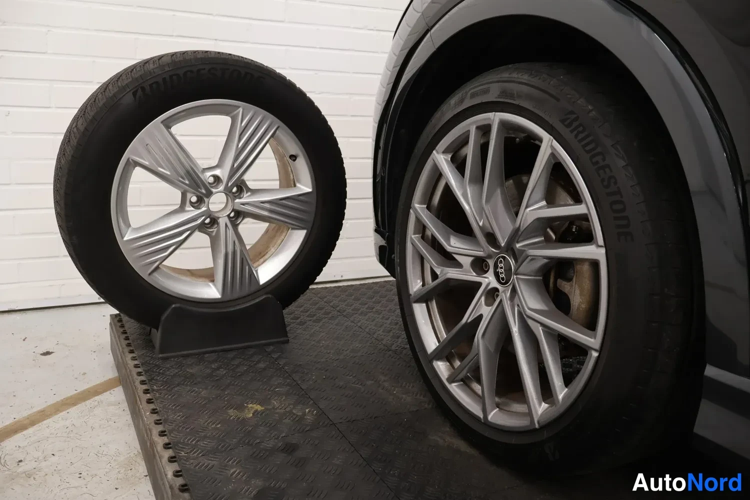 Yksityiskohta: Kaksi rengasta ja vannesuutaa: auton pyörä Bridgestone-renkaalla ja irrotettu Audi-vannesivu telineessä.