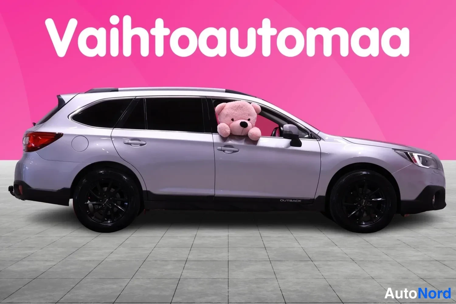 Sivulta: Full side profile of a silver Subaru Outback with black roof rails and alloy wheels. A pink teddy bear sits in the open driver's window.