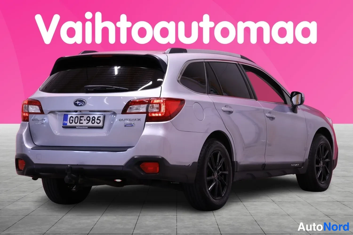 Takaviisto: Rear three-quarter view of a silver Subaru Outback with the license plate clearly visible. The rear bumper is equipped with a tow hitch and fog lights.
