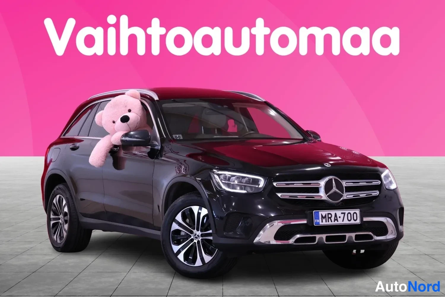 Etuviisto, nokka vasemmalle: Front three-quarter view of a black SUV against a pink background with white text 'Vaihtoautoma'. A large pink teddy bear sits in the driver's side win