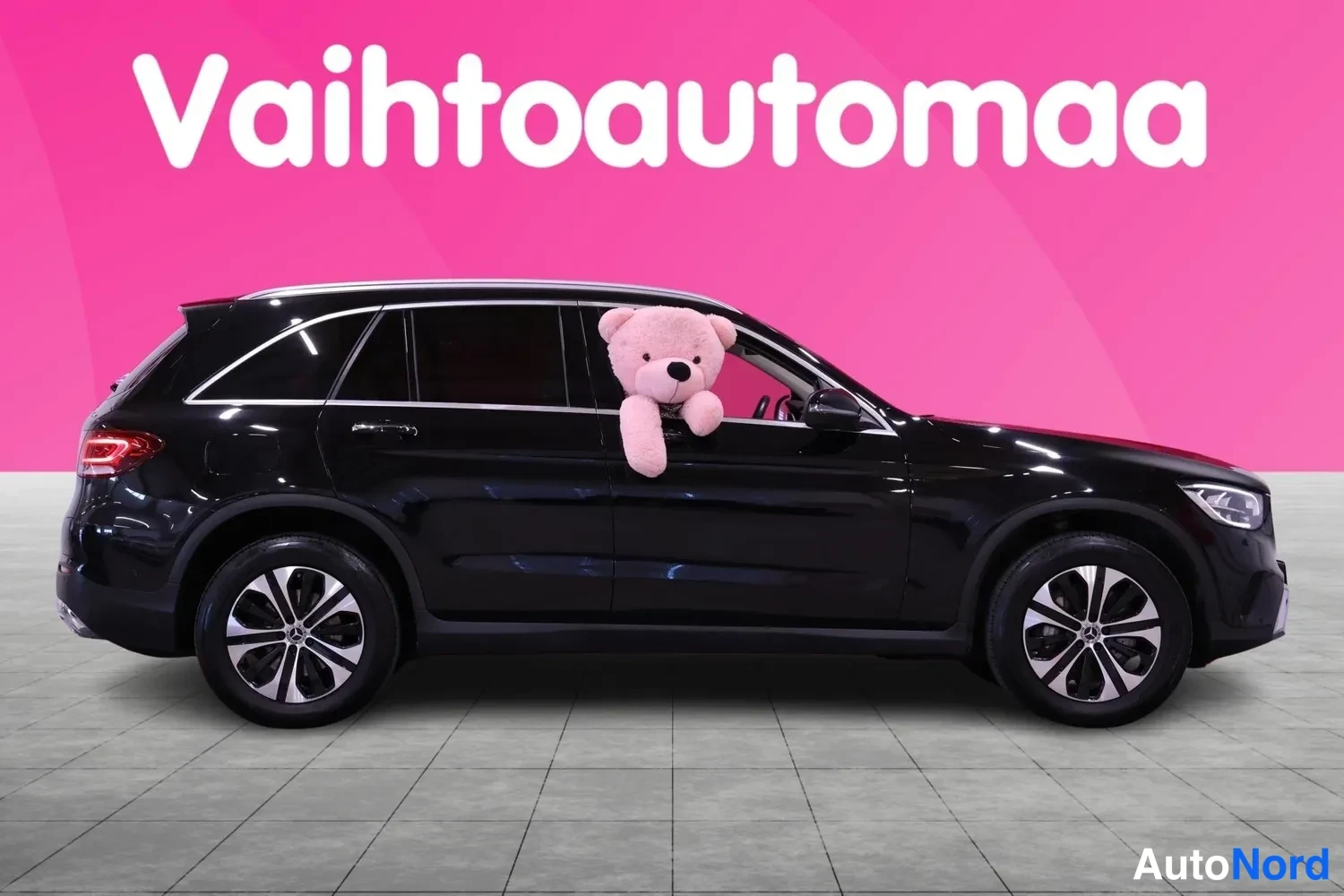 Sivulta: Side profile view of a black SUV parked indoors on a grey tiled floor against a pink wall with white text 'Vaihtoautoma'. The complete left flank of the vehicle