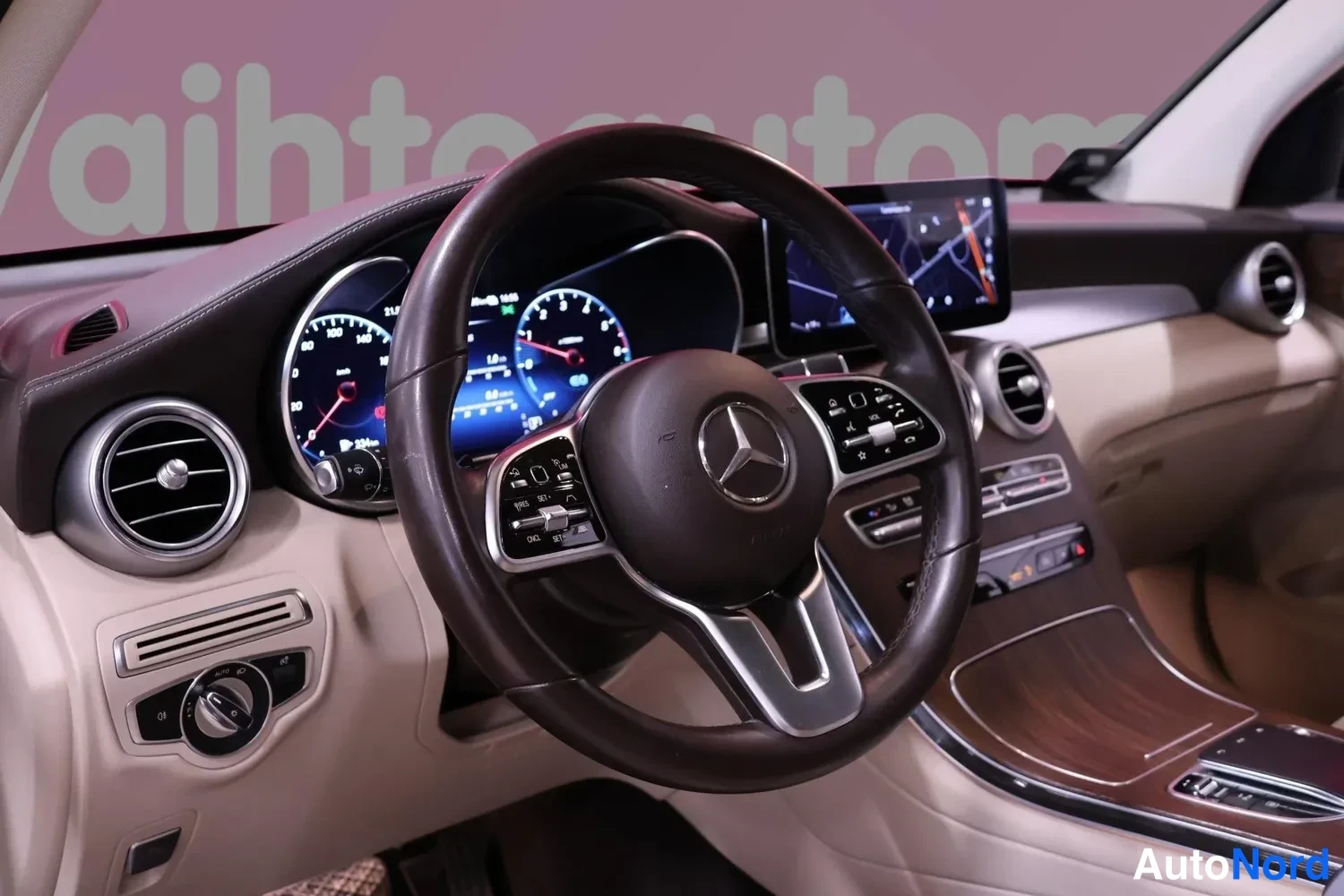 Yksityiskohta: Mercedes-Benzin ohjaamo: tumma nahkainen ohjauspyörä, näytöllä oleva mittaristo ja kosketusnäyttö navigointikartalla, beigea sisustaa ja pukuviota koristeosia.