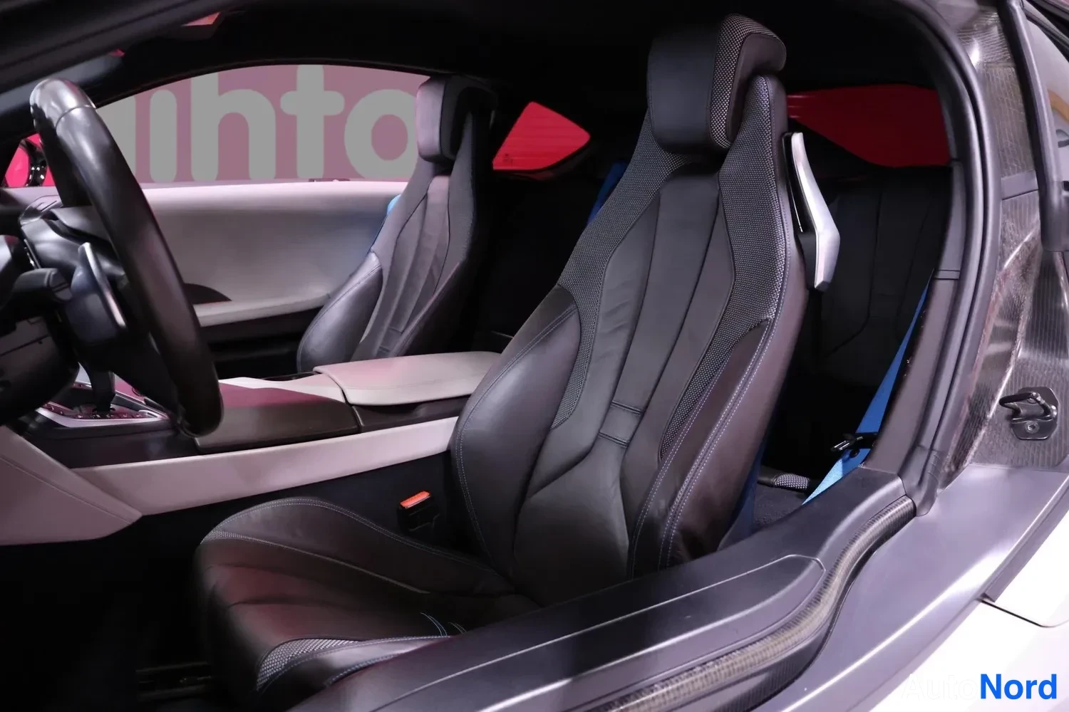 Muu: BMW i8:n sisätilat sivulta: kaksi mustaa urheiluistuinta, ohjauspyörä vasemmalla ja hiilikuitutekstuuri ovenpielessä.