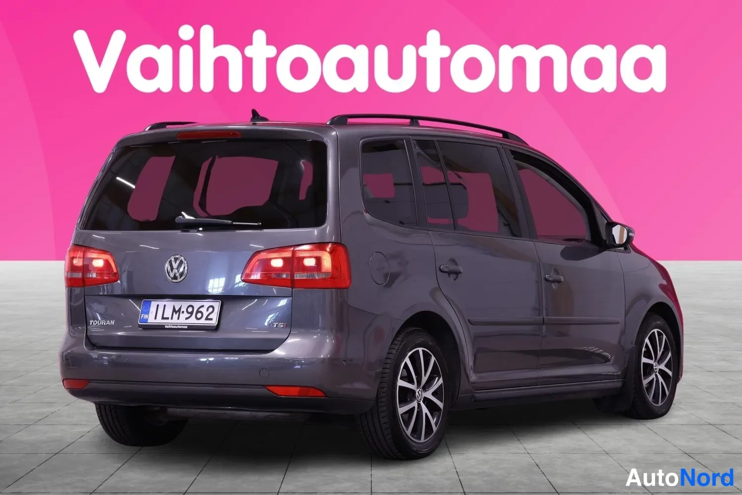 Takaviisto: Auton takaosa ja oikea kylki kuvattuna, harmaa farmariauto rekisterillä, kattokaiteet ja LED-takavalot näkyvissä, taustalla pinkki Vaihtoautomaata-logoseinä.