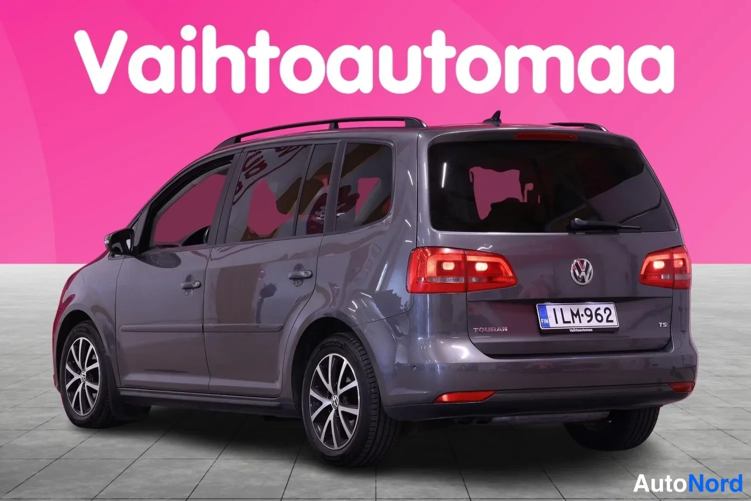 Takaviisto: Tummanharmaan tila-auton takakulmanäkymä: punaiset perävalot, VW-merkki, TOURAN- ja TSI-tekstit, rekisterikilpi sekä kattokaiteet näkyvissä.