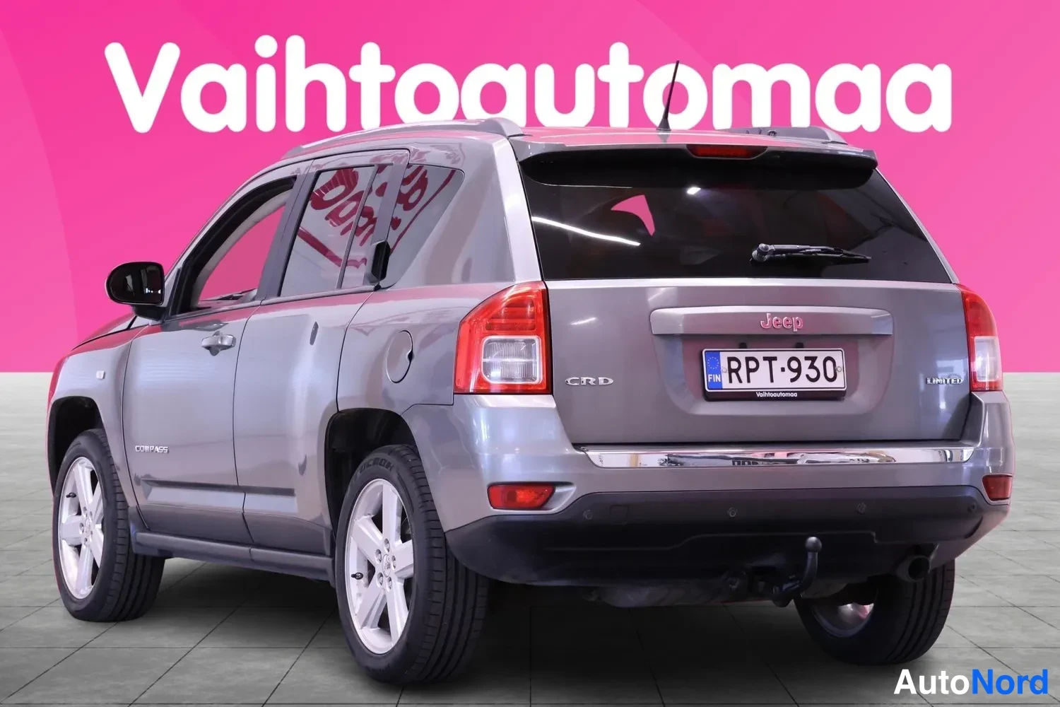 Takaviisto: Silver Jeep Compass rear and right side view showing taillights, license plate, chrome trim, 'Limited' badge on the tailgate, and tow bar. Pink background with